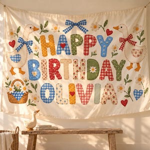 May include: A cream-colored fabric wall hanging featuring the text HAPPY BIRTHDAY OLIVIA in colorful patterned letters. The design includes two geese, a basket of daisies, small red hearts, and a blue gingham bow, all in a quilt-like style.