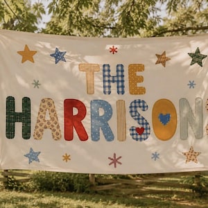 May include: A rectangular white fabric banner featuring the text THE HARRISONS in large, multi-colored, patterned fabric letters. The banner is decorated with various fabric stars and small flower shapes in yellow, blue, green, and red.