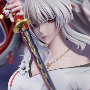 May include: A detailed anime-style figurine featuring a character with long white hair and red eyes, wearing a white garment with floral accents. The character holds a sword with a red and gold hilt. Text at the bottom reads May 2026 Patreon | Sylviana Sculpt.