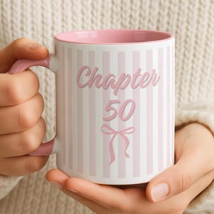 Peut inclure: Un mug en céramique avec un motif à rayures verticales rose clair et blanc. Le texte "Chapter 50" est imprimé en rose, centré sur le devant, au-dessus d'un petit nœud rose. Le mug possède une anse rose unie et un bord intérieur rose.
