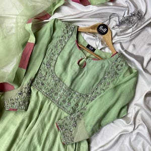 May include: A pale green dress with detailed silver embroidery on the neckline and sleeves. The dress features long sleeves and a flowing skirt. A matching light green scarf with pink trim is visible. The dress is on a wooden hanger.