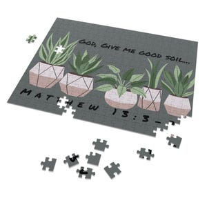 May include: A partially assembled jigsaw puzzle with a gray background. The puzzle features illustrations of potted plants with the text "GOD, GIVE ME GOOD SOIL..." and "MATTHEW 13:3-". Several puzzle pieces are scattered around the main puzzle.