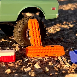 May include: A miniature light green truck with a black tire, two bright orange traction boards, a small red and white cooler, and a blue plastic fuel canister. The items are arranged together on a textured, rocky surface in natural lighting.