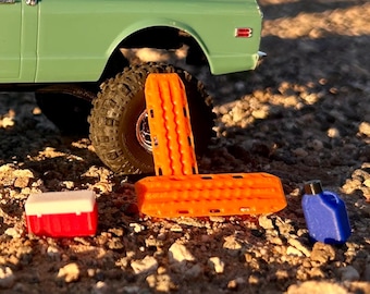 May include: A miniature light green truck with a black tire, two bright orange traction boards, a small red and white cooler, and a blue plastic fuel canister. The items are arranged together on a textured, rocky surface in natural lighting.