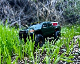 May include: A dark green Toyota Tacoma scale model pickup truck featuring a black grille with white Toyota lettering, a roof-mounted light bar with yellow lenses, bronze-colored wheels, and a red storage box mounted in the truck bed.