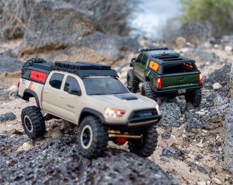 May include: Two remote-controlled Toyota Tacoma pickup trucks navigating rocky terrain. The front truck is tan with a black roof rack and red side panels, while the rear truck is dark green with a black bed cap. Both feature off-road tires.