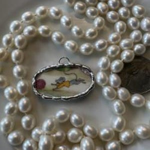 Puede incluir: Un colgante de plata con un diseño de ratón pintado a mano, ensartado en una doble hebra de cuentas de perlas blancas. El colgante presenta un ratón con un atuendo amarillo sosteniendo una pelota roja. Las perlas están sobre un plato blanco.