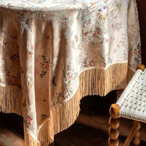 May include: A beige round tablecloth with a floral and butterfly pattern. The tablecloth has a fringe trim and is draped over a table. The floral design includes red, pink, and blue flowers.