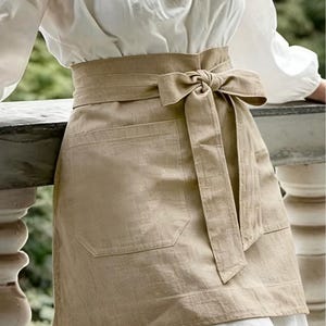 May include: A beige waist apron featuring a wide waistband that ties in a bow at the front. The apron includes a large, rectangular patch pocket on the front side, constructed from a textured fabric with visible weave details and a straight hemline.