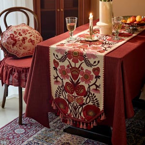 May include: A rectangular table runner featuring a cream-coloured centre with a symmetrical floral pattern in shades of red, pink, and dark brown. The runner is finished with a decorative fringe of tassels along the bottom edge and is placed on a solid dark red tablecloth.