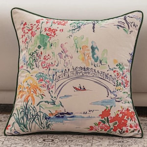 Op de afbeelding: Vierkant decoratief kussen met een kleurrijk aquarel schilderij van een brug, bomen en bloemen. Het kussen heeft een crèmekleurige achtergrond met een groene rand. Het kunstwerk heeft rode, oranje, groene en blauwe tinten.