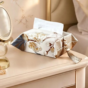 Pode incluir: Uma capa retangular para caixa de lenços feita de tecido de cetim na cor champanhe com um padrão floral bordado em tons de branco, dourado e azul. A capa inclui uma borla decorativa a condizer presa a um lado com um detalhe de conta.
