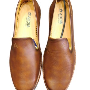 Pu&ograve; includere: Un paio di scarpe slip-on in pelle marrone. Le scarpe hanno la punta arrotondata e la suola cucita. L'interno &egrave; di colore marrone chiaro, con il marchio "ADDIS" visibile. Le scarpe sono esposte su sfondo bianco.