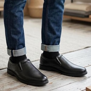 Op de afbeelding: Zwarte leren instappers met een getextureerde afwerking. De schoenen worden gecombineerd met een donkerblauwe jeans met opgerolde manchetten en zwarte sokken. De schoenen hebben een laag profiel en een eenvoudig ontwerp, geschikt voor casual of semi-formeel gebruik.