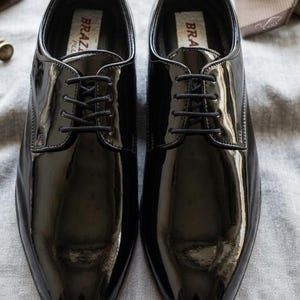 May include: A pair of shiny black dress shoes with laces, displayed on a gray surface. The shoes have a glossy finish and a classic design, suitable for formal occasions. The brand name "BRAZ" is visible on the inner label.