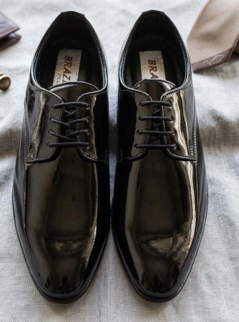 May include: A pair of shiny black dress shoes with laces, displayed on a gray surface. The shoes have a glossy finish and a classic design, suitable for formal occasions. The brand name "BRAZ" is visible on the inner label.