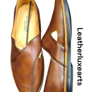 Puede incluir: Dos zapatos sin cordones de cuero marr&oacute;n. Los zapatos tienen un dise&ntilde;o cosido y una suela negra con costuras amarillas. El interior del zapato es de color beige claro. El texto "LAJJIS MADE IN INDIA" es visible.
