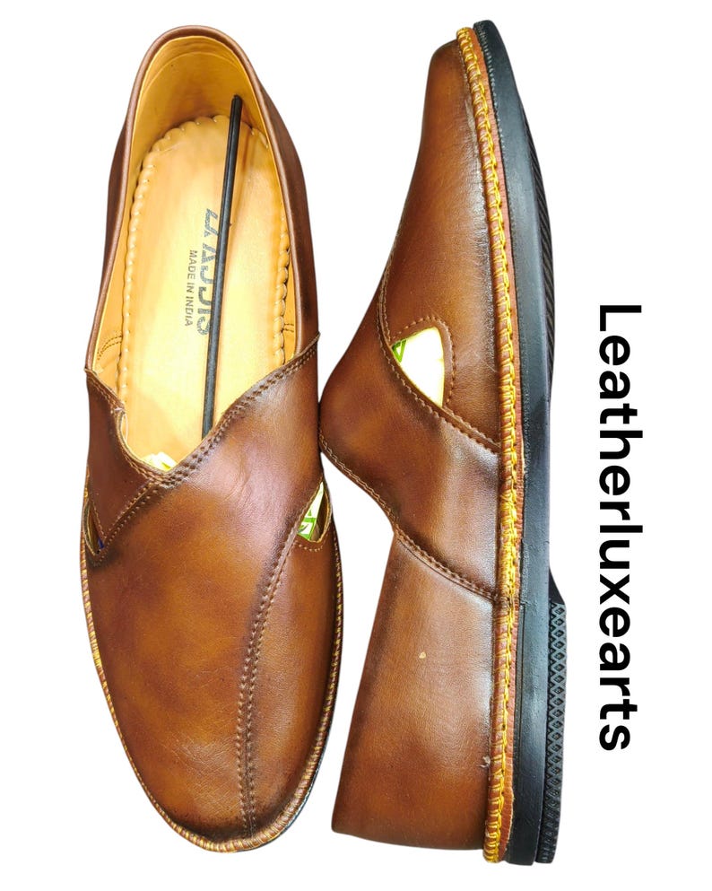 Puede incluir: Dos zapatos sin cordones de cuero marr&oacute;n. Los zapatos tienen un dise&ntilde;o cosido y una suela negra con costuras amarillas. El interior del zapato es de color beige claro. El texto "LAJJIS MADE IN INDIA" es visible.