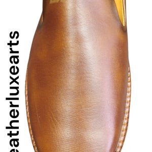 Pu&ograve; includere: Primo piano di una scarpa in pelle marrone. La scarpa ha una punta arrotondata e una suola cucita. La tomaia &egrave; liscia e lucida, con un logo sottile in alto. Il testo "Leatherluxearts" &egrave; stampato verticalmente a sinistra.