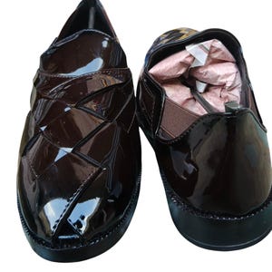 May include: A pair of dark brown patent leather slip-on shoes with a woven-style upper design, black soles, and elastic side panels. The interior of the right shoe contains pink paper stuffing. The text "Leatherluxearts" appears below the shoes.