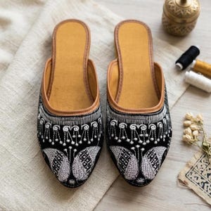 May include: A pair of black slip-on mules featuring intricate white embroidery on the toe area. The shoes have a pointed toe, a tan interior lining, and a tan trim along the opening. The embroidery displays a repeating pattern of stylized shapes.