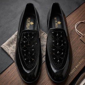 May include: Pair of black patent leather loafers with a velvet vamp featuring a geometric pattern of studs. The shoes have a glossy finish and are displayed on a wooden surface. The brand name "BLUE HILLS" is visible on the insole.