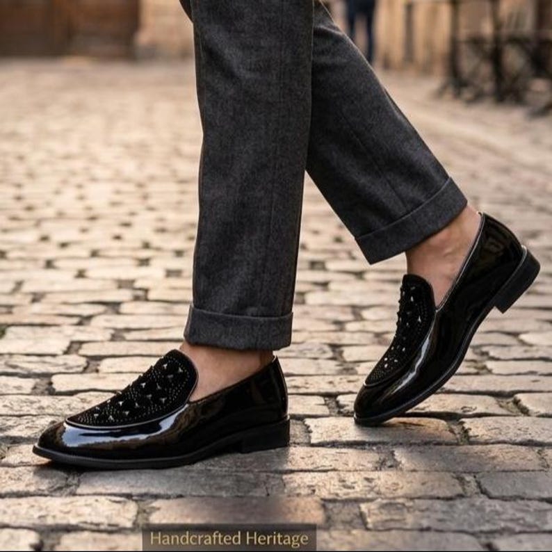 May include: Black patent leather loafers with studded detailing on the vamp. The shoes have a glossy finish and a low heel. The image also shows gray trousers and cobblestone pavement. The text "Handcrafted Heritage" is at the bottom of the image.