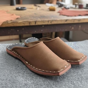 Op de afbeelding: Een paar bruine leren slippers met een vierkant teenontwerp. De slippers hebben een donkerbruin interieur en een lichtbruine zool met witte stiksels rond de randen. Ze liggen op een grijze ondergrond.