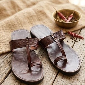 May include: A pair of dark brown leather sandals featuring a toe-post design, a wide strap across the instep with an embossed pattern, and two small metal rivets. The sandals are displayed on a wooden surface with the text Leatherluxearts below.
