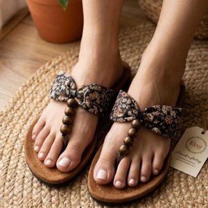 May include: A pair of thong sandals featuring tan soles and a T-strap design adorned with three dark brown wooden beads. The upper strap is made of black fabric with a multi-colored floral pattern tied into a bow shape over the foot.