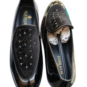 May include: Pair of black patent leather loafers with a velvet vamp adorned with silver-tone studs. The shoes have a glossy finish and a classic slip-on design. The brand name "Blue Hills" is visible on the insole.