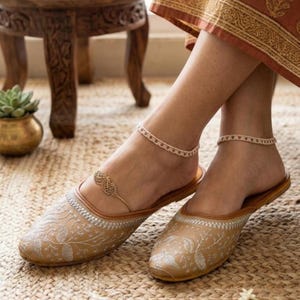 May include: A pair of tan slip-on shoes featuring intricate white floral embroidery and a pointed toe, paired with two delicate gold-toned anklets, one with a small filigree leaf-shaped charm and the other with a simple beaded chain design.