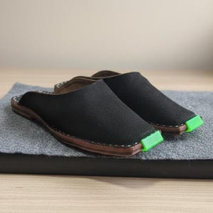 Può includere: Un paio di scarpe nere slip-on con un design a punta quadrata. Le scarpe hanno una suola marrone e un accento verde sulla punta. Le scarpe sono posizionate su un tappetino grigio.