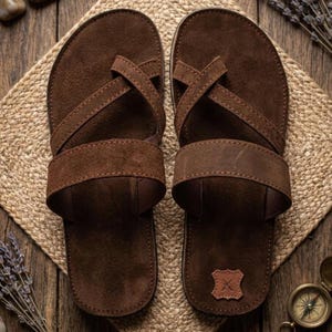 Puede incluir: Un par de sandalias de ante marrón con diseño de tiras. Las sandalias tienen una tira ancha en la parte superior del pie y una tira más pequeña que va entre los dedos. Las sandalias están colocadas sobre una estera tejida.