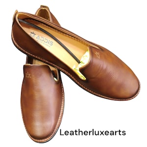 Pu&ograve; includere: Mocassini in pelle marrone dal design classico. Le scarpe hanno una finitura liscia e lucida e una punta arrotondata. Il marchio "ADDIS" &egrave; visibile sul lato. Il testo "Leatherluxearts" &egrave; in basso.