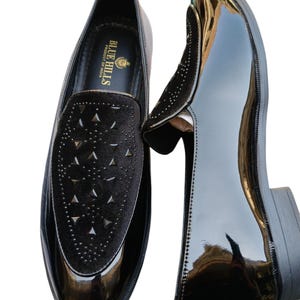 May include: A pair of black patent leather loafers with a velvet vamp featuring triangle cutouts. The shoes have a glossy finish and a low heel. The brand "BLUE HILLS PRODUCT OF INDIA" is visible inside the shoe. The text "Leatherluxearts" is at the bottom.