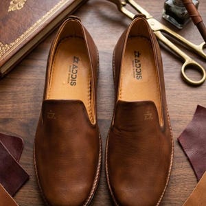 Pu&ograve; includere: Un paio di mocassini in pelle marrone con il marchio "ADDIS" visibile all'interno. Le scarpe sono esposte su una superficie di legno, insieme a campioni di pelle, un libro d'epoca e strumenti antichi.