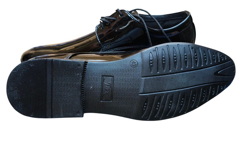 May include: Pair of glossy black dress shoes with laces. One shoe is upright, the other is upside down, revealing the textured sole. The shoes have a classic design, suitable for formal occasions.