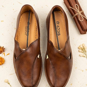 Puede incluir: Un par de zapatos sin cordones de cuero marr&oacute;n con un dise&ntilde;o recortado &uacute;nico. Los zapatos tienen una costura y la marca "ADDIS" es visible. Los zapatos est&aacute;n hechos en India. Un art&iacute;culo de cuero enrollado atado con hilo est&aacute; en el fondo.