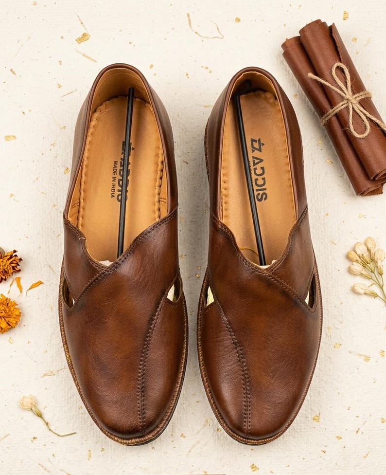 Puede incluir: Un par de zapatos sin cordones de cuero marr&oacute;n con un dise&ntilde;o recortado &uacute;nico. Los zapatos tienen una costura y la marca "ADDIS" es visible. Los zapatos est&aacute;n hechos en India. Un art&iacute;culo de cuero enrollado atado con hilo est&aacute; en el fondo.