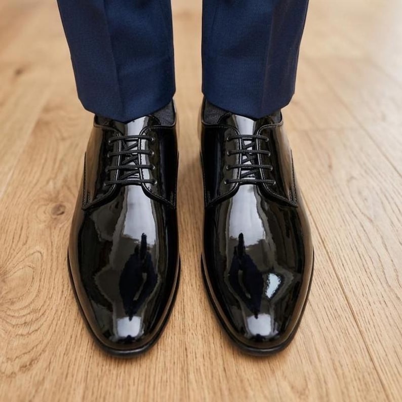 May include: Close-up of a pair of shiny black leather dress shoes with laces. The shoes are highly polished, reflecting the light. The shoes are paired with navy blue trousers, suggesting formal wear.
