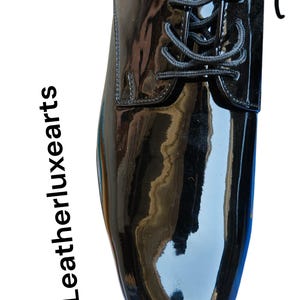May include: A glossy black dress shoe with a lace-up design. The shoe has a patent leather finish, reflecting light. The text "Leatherluxearts" is printed vertically on the left side of the image.