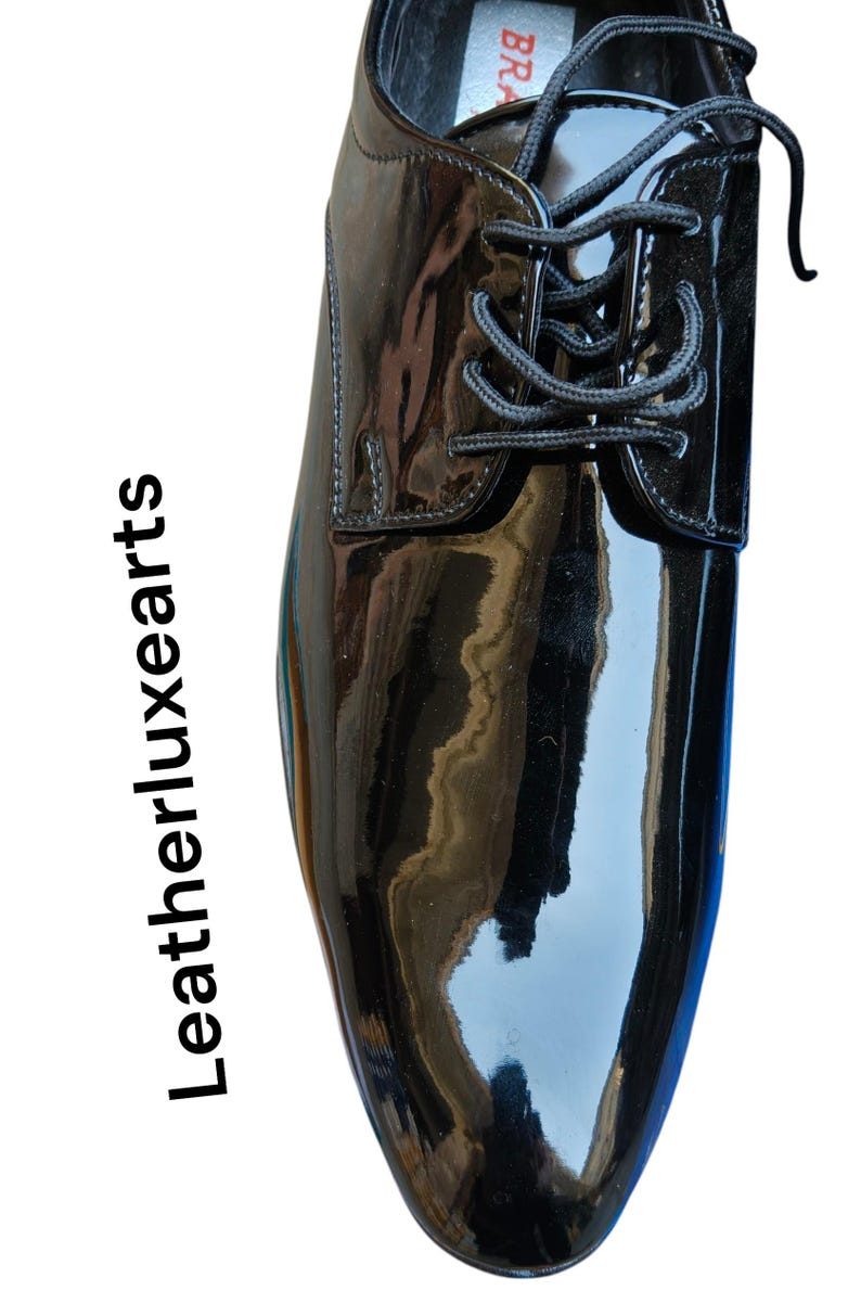 May include: A glossy black dress shoe with a lace-up design. The shoe has a patent leather finish, reflecting light. The text "Leatherluxearts" is printed vertically on the left side of the image.