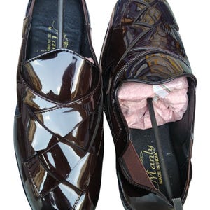May include: A pair of dark brown loafers with a high-gloss patent finish. The upper features a woven-style overlapping strap design. The interior of the shoe is black with the text "Manly" and "MADE IN INDIA" printed on the insole.