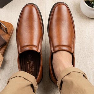 Puede incluir: Un par de zapatos sin cordones de cuero marrón, con punta redondeada y suelas negras. Los zapatos se muestran desde arriba, mostrando su textura suave y diseño. La marca "Doctor + Orthotics.walk" es visible en la plantilla.
