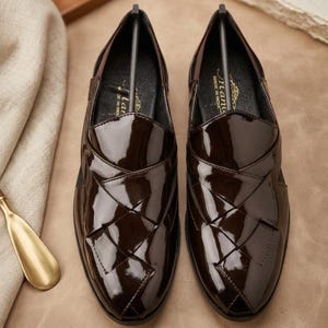 May include: A pair of dark brown patent leather loafers featuring a woven-style upper design with a glossy finish. The interior of each shoe includes a black insole with the text "Manu" and "MADE IN INDIA" printed in gold lettering.