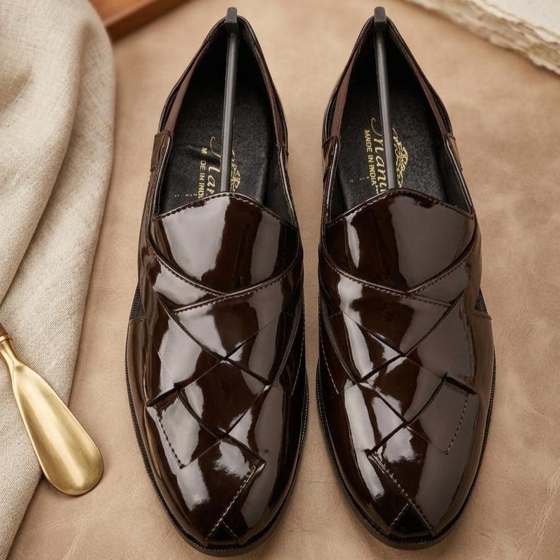May include: A pair of dark brown patent leather loafers featuring a woven-style upper design with a glossy finish. The interior of each shoe includes a black insole with the text "Manu" and "MADE IN INDIA" printed in gold lettering.