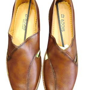 Puede incluir: Un par de zapatos sin cordones de cuero marr&oacute;n con un dise&ntilde;o cosido y detalles recortados. Los zapatos tienen un interior marr&oacute;n claro y el texto "ADDIS MADE IN INDIA" es visible en el interior. La marca "Leatherluxearts" est&aacute; en la parte superior.