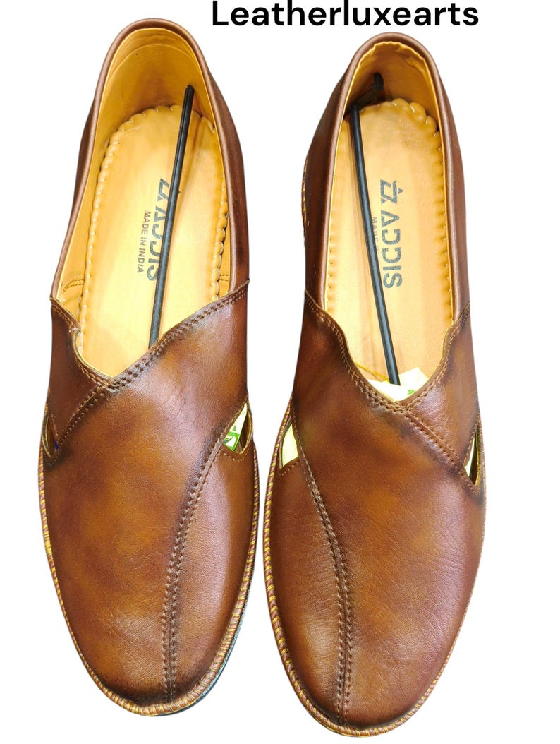 Puede incluir: Un par de zapatos sin cordones de cuero marr&oacute;n con un dise&ntilde;o cosido y detalles recortados. Los zapatos tienen un interior marr&oacute;n claro y el texto "ADDIS MADE IN INDIA" es visible en el interior. La marca "Leatherluxearts" est&aacute; en la parte superior.