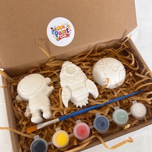 May include: A space-themed paint-your-own kit in a cardboard box. The kit contains a white astronaut, rocket, and planet, a paintbrush, and six paint pots. The box also includes brown paper filler and a sticker that says "Mini Paint Club".